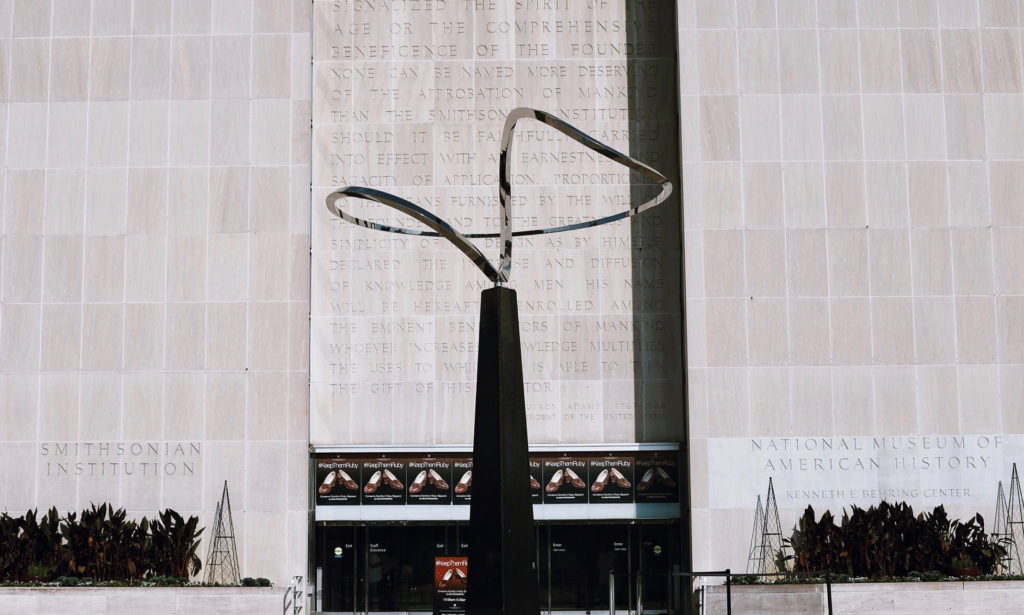 A tall, modern sculpture stands in front of a large building with engraved text. Below are glass doors and signage for the Smithsonian National Museum of American History. Plants line the entrance.
