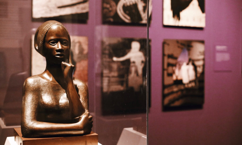 A bronze sculpture of a thoughtful woman is displayed in a museum, set against a background of blurred paintings on a purple wall. The sculptures serene expression adds to the contemplative atmosphere.