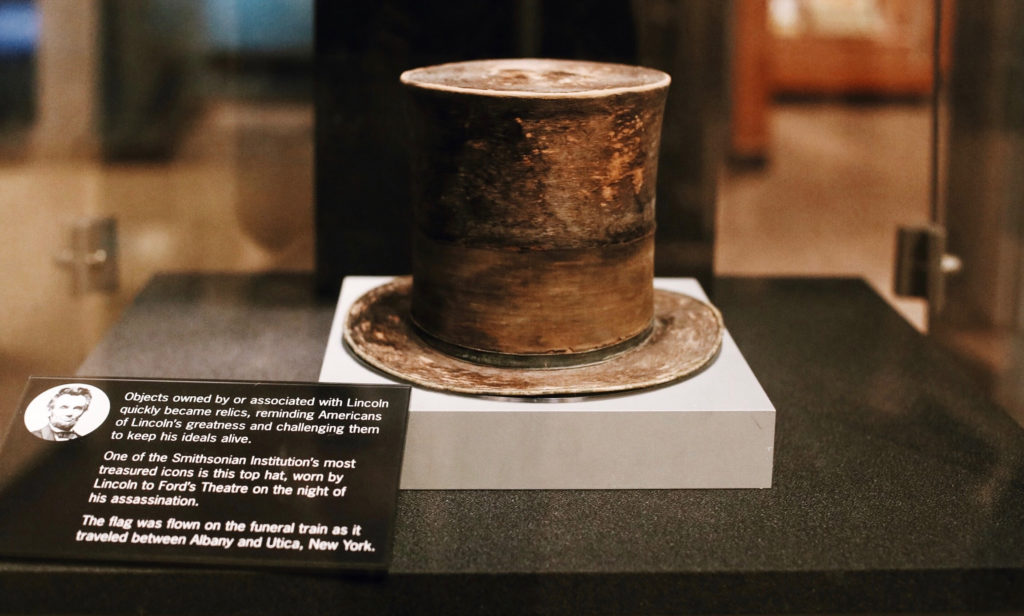 A historic top hat displayed in a clear case with an informational plaque. The plaque mentions its association with Lincoln and highlights its significance as a relic from Fords Theatre on the night of his assassination.