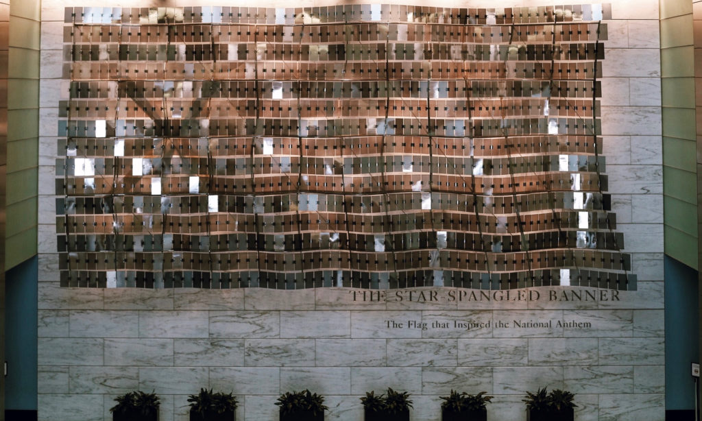 A wall display titled The Star-Spangled Banner features reflective metal panels arranged in a wave pattern. Below is a caption, The Flag that Inspired the National Anthem. Seven small potted plants line the floor in front.