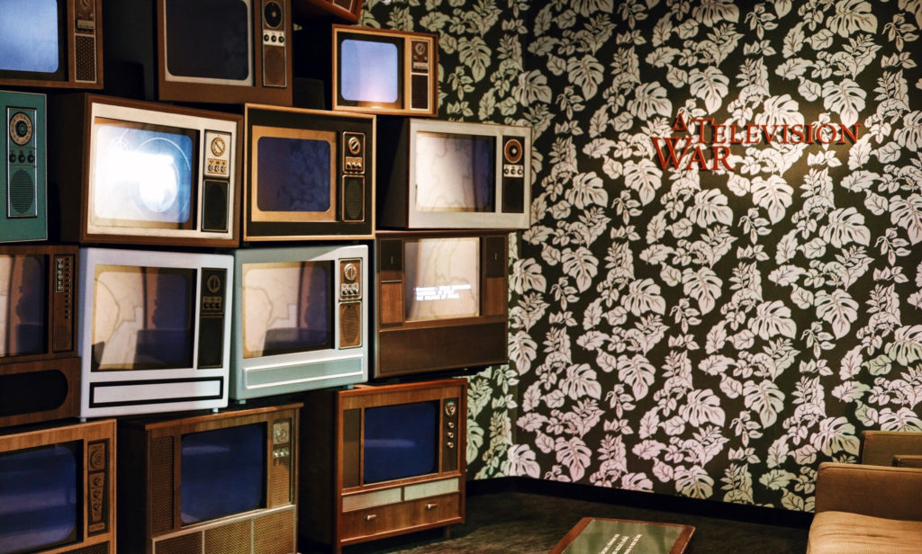 A wall with vintage televisions stacked in a mosaic design next to a floral-patterned wallpaper. A small sign reads TELEVISION WAR. The scene has a retro aesthetic, with muted brown and green tones.