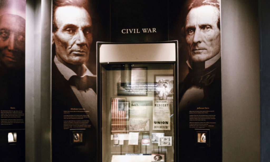 Museum exhibit on the Civil War, featuring a display case with artifacts such as documents and flags. Large portraits of historical figures are on the surrounding walls.