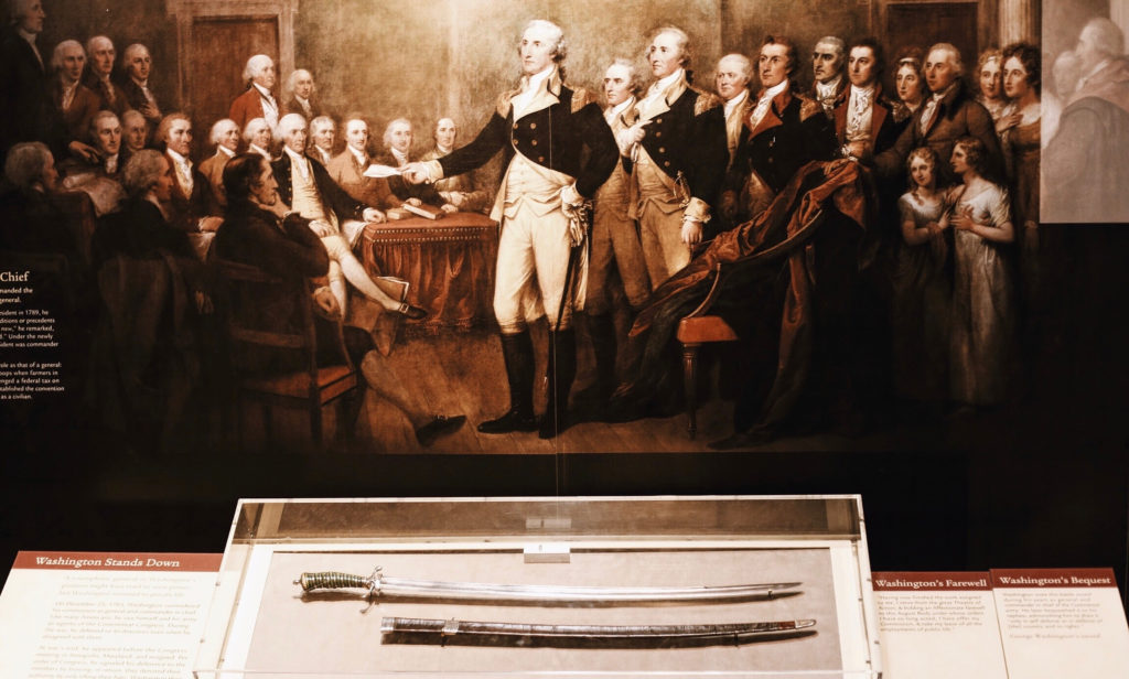 A historical painting depicts George Washington standing in a formal room, surrounded by several men in 18th-century attire. Below the painting is a display case containing a sword and its sheath. Informative plaques are placed in front of the case.
