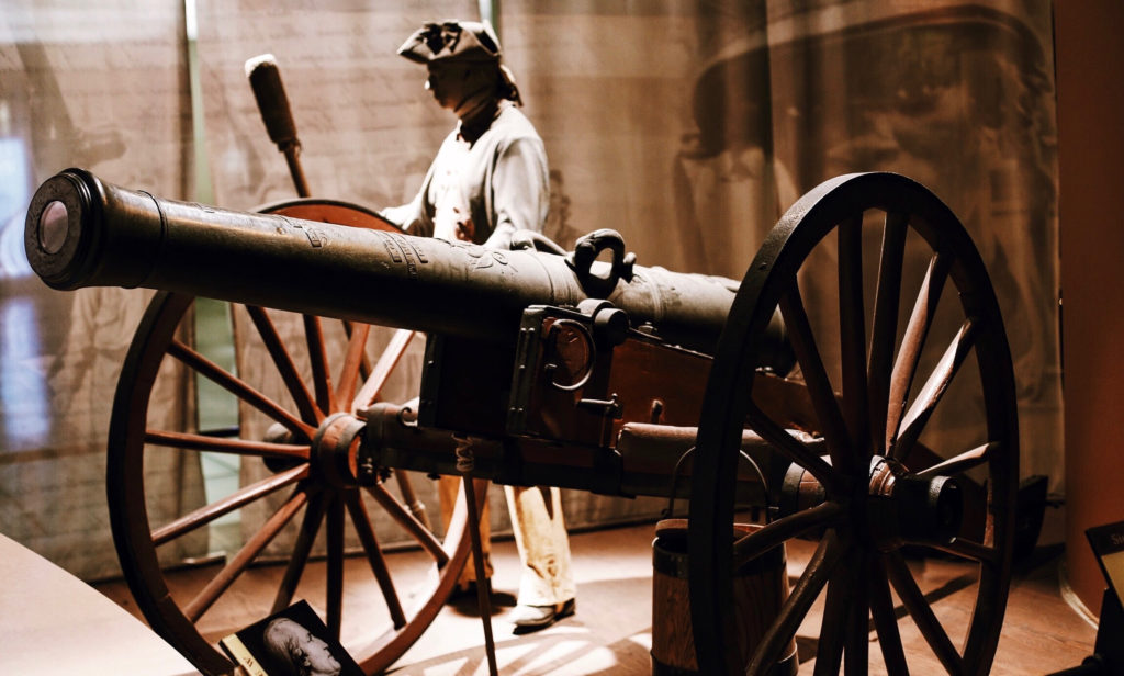 A historical museum display featuring a traditional cannon on wooden wheels with metal accents. Behind it stands a mannequin dressed in 18th-century soldier attire, creating an immersive, period-appropriate setting.