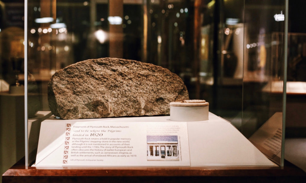 A fragment of Plymouth Rock displayed in a glass case at a museum. The rock is surrounded by informational text about its historical significance and the Pilgrims. Nearby exhibits are slightly visible in the background.