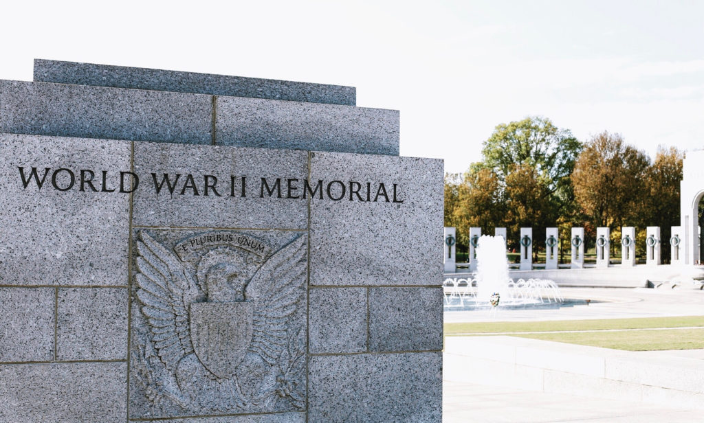 Image of the World War II Memorial with an engraved stone featuring an eagle emblem and the words World War II Memorial. In the background, a fountain and a semi-circle of pillars are visible under a clear sky.