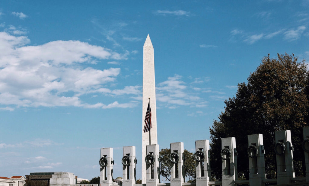 A tall, white obelisk stands against a blue sky with clouds, surrounded by green trees and a row of stone monuments. An American flag is displayed in front of the obelisk.