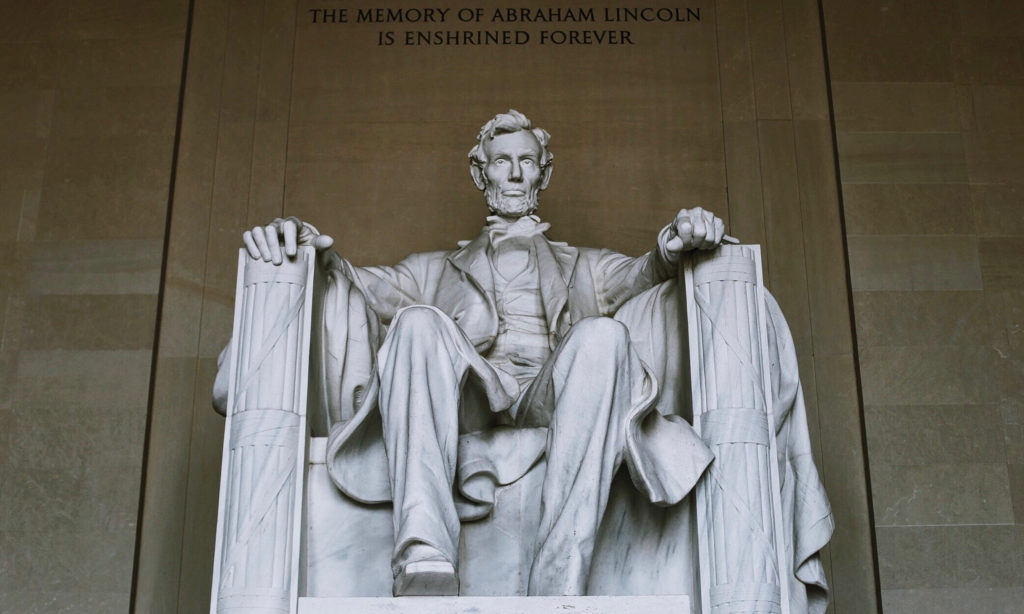 The image shows the statue of Abraham Lincoln seated in the Lincoln Memorial. The inscription above reads, The memory of Abraham Lincoln is enshrined forever. The statue is detailed, capturing his thoughtful expression.