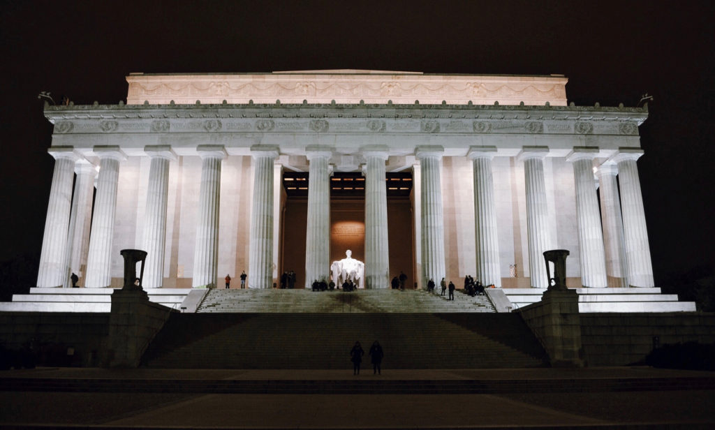 The image shows the Lincoln Memorial at night, illuminated by lights. The grand structure features large columns and steps leading up to the entrance, where the statue of Abraham Lincoln is visible in the center.