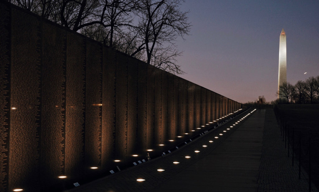 The image shows the Vietnam Veterans Memorial at dusk, with engraved names illuminated by ground lights. In the background, the Washington Monument stands tall against the evening sky. Bare trees frame the memorials reflective surface.