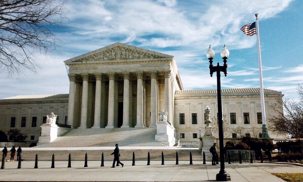 The image shows the United States Supreme Court building, characterized by its grand neoclassical architecture with tall columns and a triangular pediment. An American flag stands to the right, and a few people walk in front of the building.