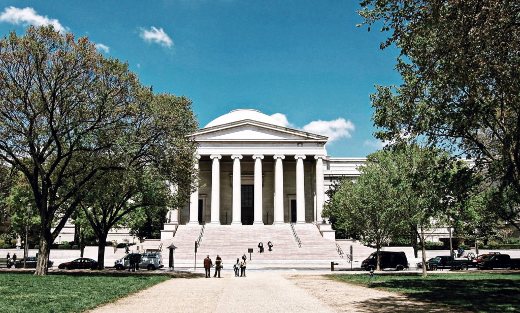 A grand neoclassical building with tall columns and a dome, surrounded by trees, under a bright blue sky. People walk along a path leading to the entrance, with cars parked on either side.