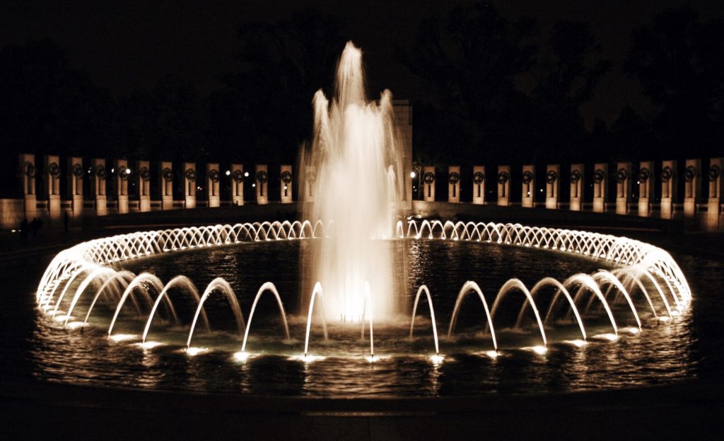 A nighttime view of a circular fountain with illuminated jets of water and a tall central spout. The fountain is surrounded by a ring of columns and soft lighting, set against a dark, tree-lined background.