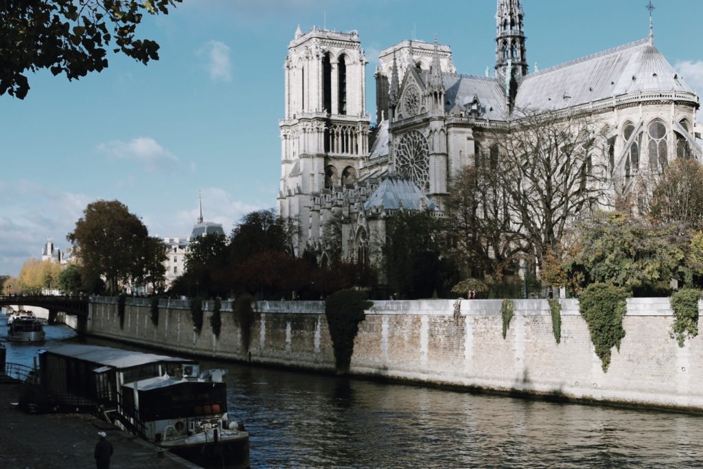 A view of Notre-Dame Cathedral in Paris, with its iconic gothic architecture. The cathedral is situated next to the Seine River, lined with trees. A boat is moored along the riverbank under a clear blue sky.