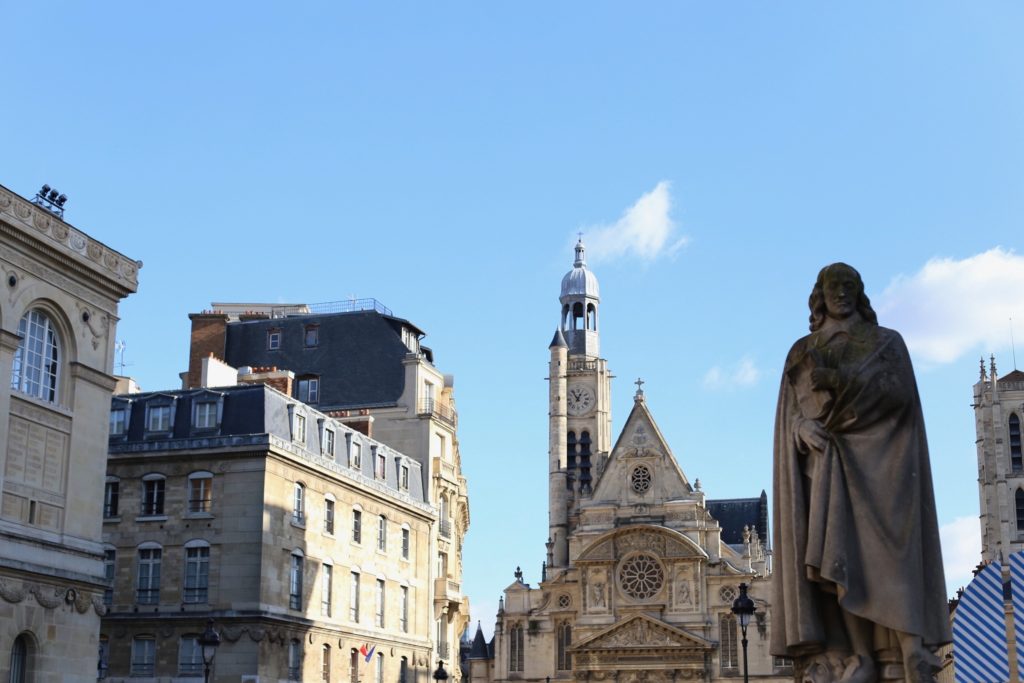Statue in front of historic buildings under a bright blue sky. A church with a prominent clock tower is visible in the background, framed by classic European architecture.