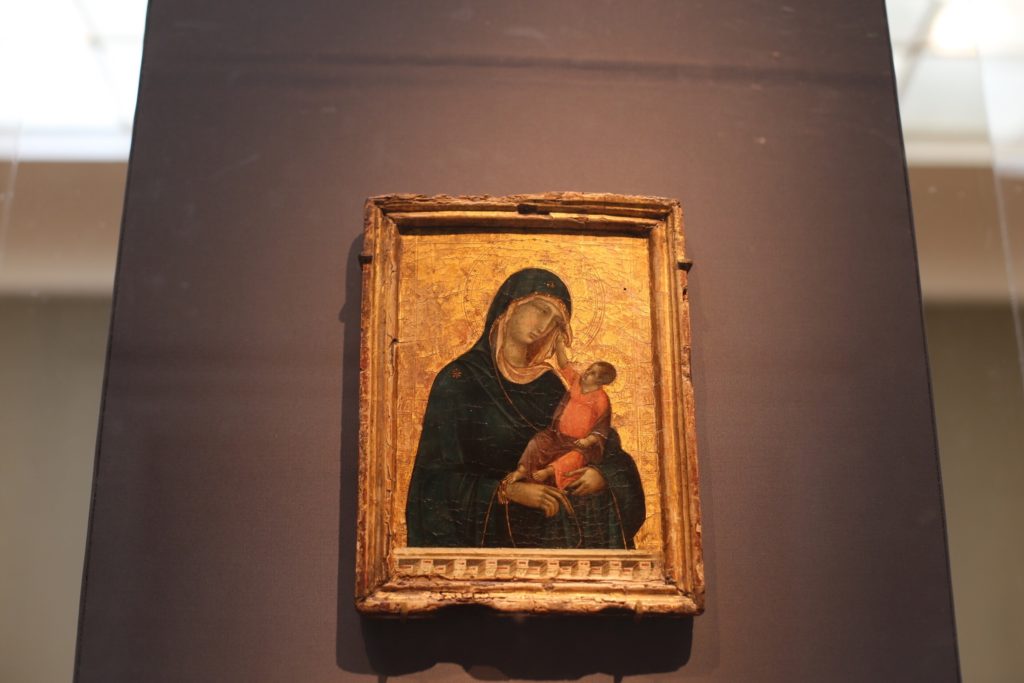 A small, framed religious painting depicting a woman in a dark robe holding a child. The artwork has a golden background and is displayed against a neutral wall.
