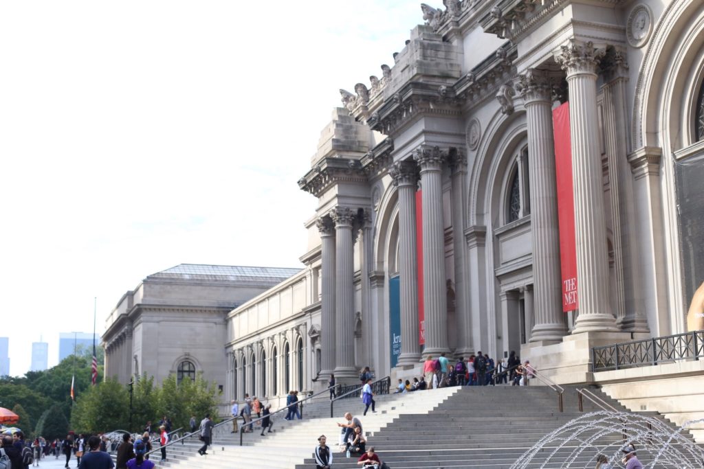 The image shows the front entrance of a large museum, featuring grand columns and wide stone steps. People are gathered on the steps and near the entrance. Trees and skyscrapers are visible in the background.