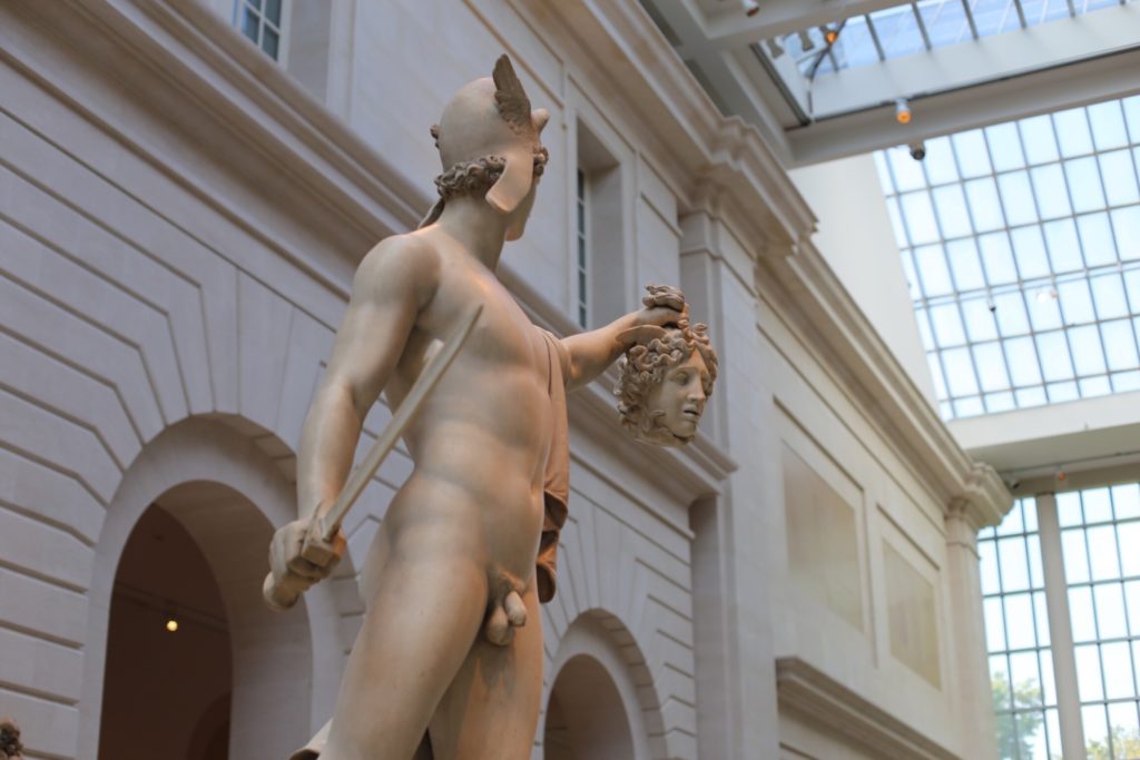 A marble statue depicting a classical figure holding a sword in one hand and the severed head of Medusa in the other. The sculpture is set in a museum interior with a high glass ceiling.