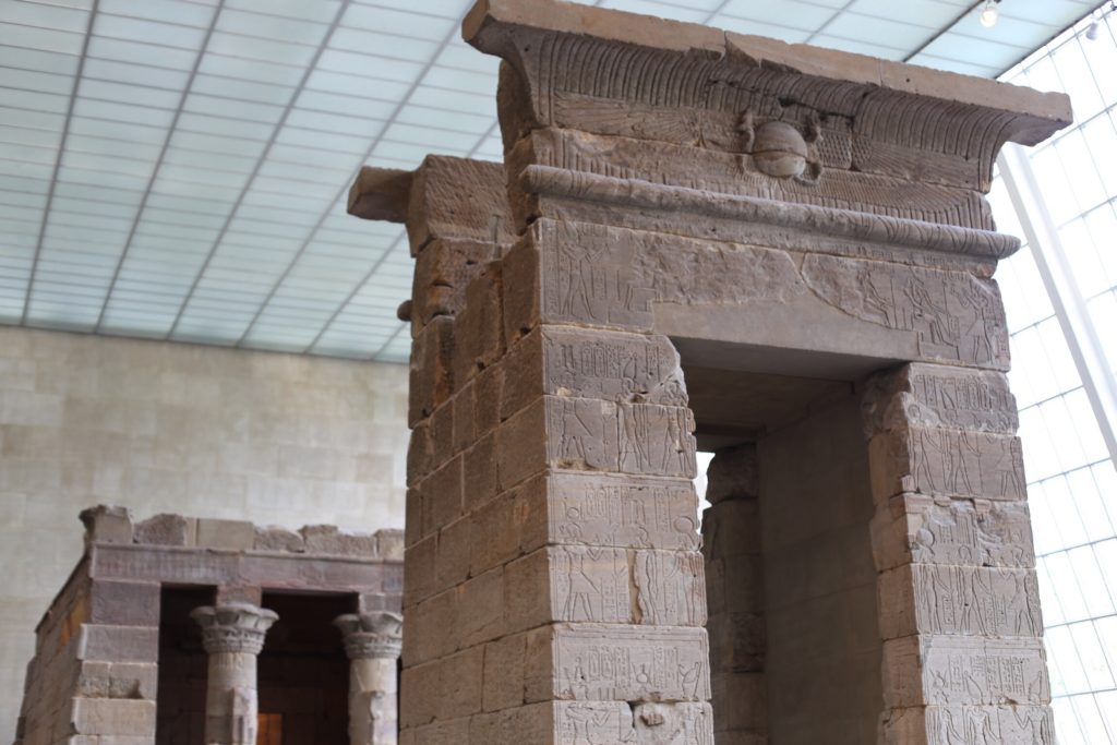 Ancient stone temple interior with detailed carvings on pillars and walls, featuring hieroglyphic inscriptions and ornate designs. The structure is illuminated by natural light from the large, grid-panel windows above.