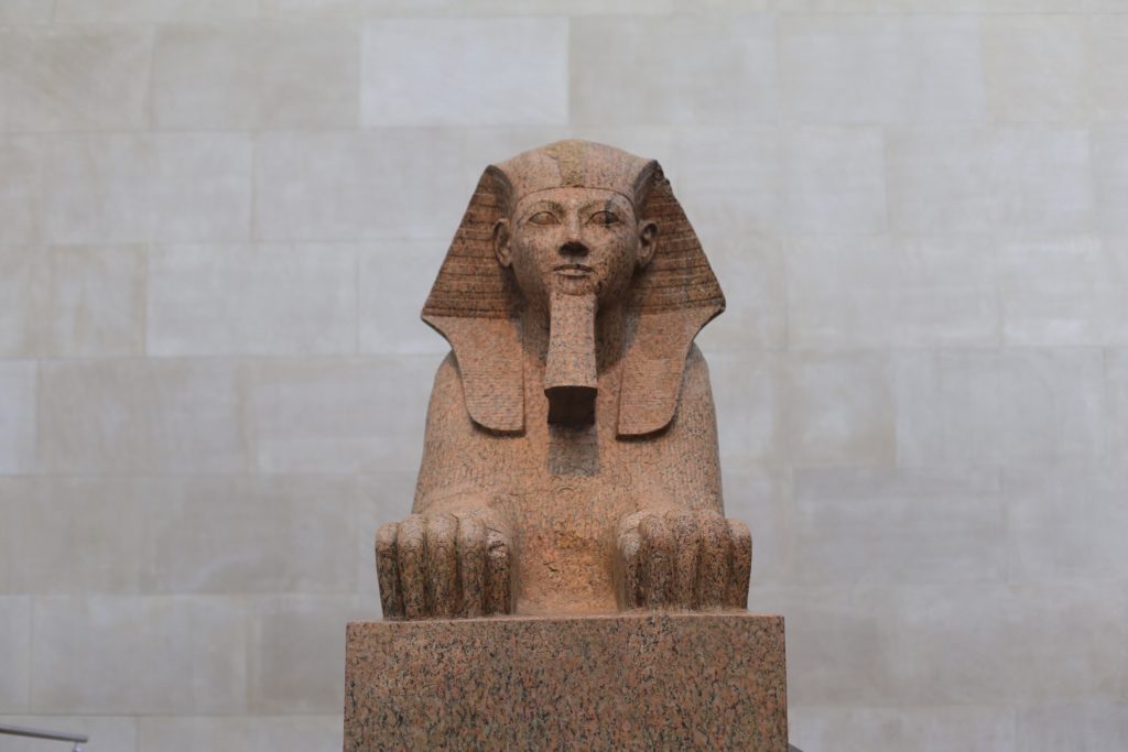 A large stone statue of an ancient Egyptian sphinx sits in a museum setting. The sphinx features a human head with a headdress and the body of a lion, intricately carved from red granite. The background is a plain, light stone wall.
