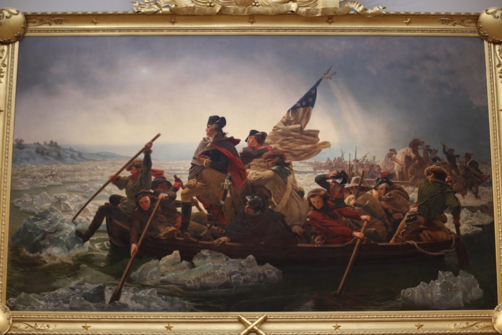 A historical painting depicting a group of men in a boat crossing an icy river. The leader, draped in a cloak, stands prominently, holding a flag. The scene conveys urgency and determination amidst a frozen landscape.