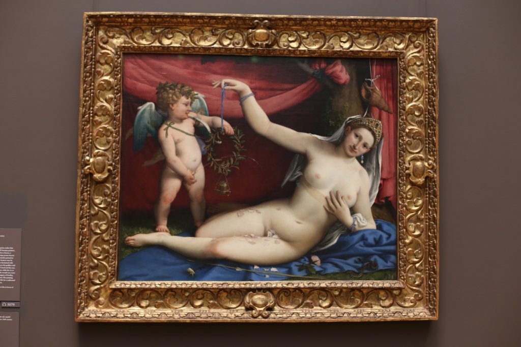 A framed classical painting depicting a reclining nude woman with a cherub. The woman lies on a blue drape, reaching towards the cherub holding a wreath. The background features a red curtain and detailed ornamentation on the gold frame.