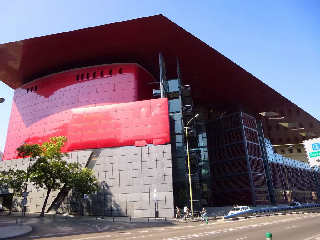 Modern building with a vibrant red upper section and large overhanging roof. The lower facade features grey and black panels. Located on a corner with a few people and a car nearby. Clear blue sky in the background.