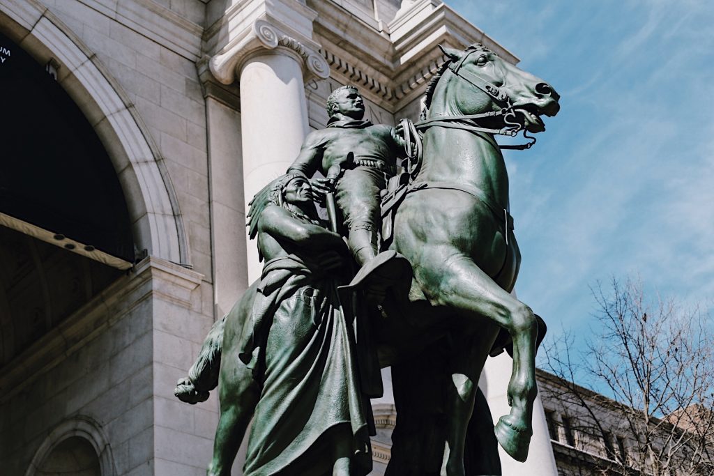 A bronze equestrian statue showcases a rider in historical military attire, mounted on a horse with dynamic posture. The statue is set against a backdrop of a grand building with pillars and an archway, under a clear blue sky.