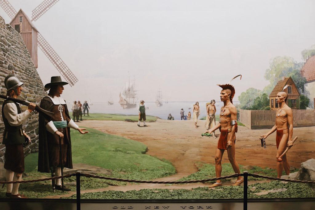 A diorama depicting early colonial New York. On the left, two European settlers in traditional attire, near a windmill. On the right, two Indigenous figures with tools. Ships and more settlers can be seen in the background.