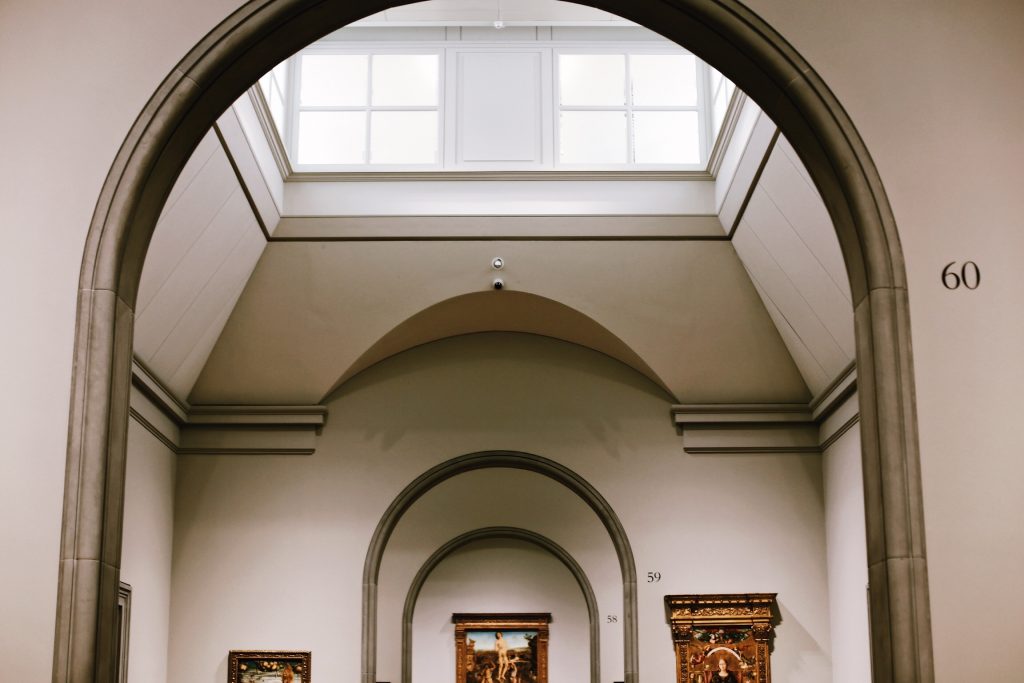 Interior of an art gallery displaying framed paintings, with a large arched doorway and a skylight. Numbers 59 and 60 are visible on the walls near the paintings. The ceiling is high and decorated with additional arches.