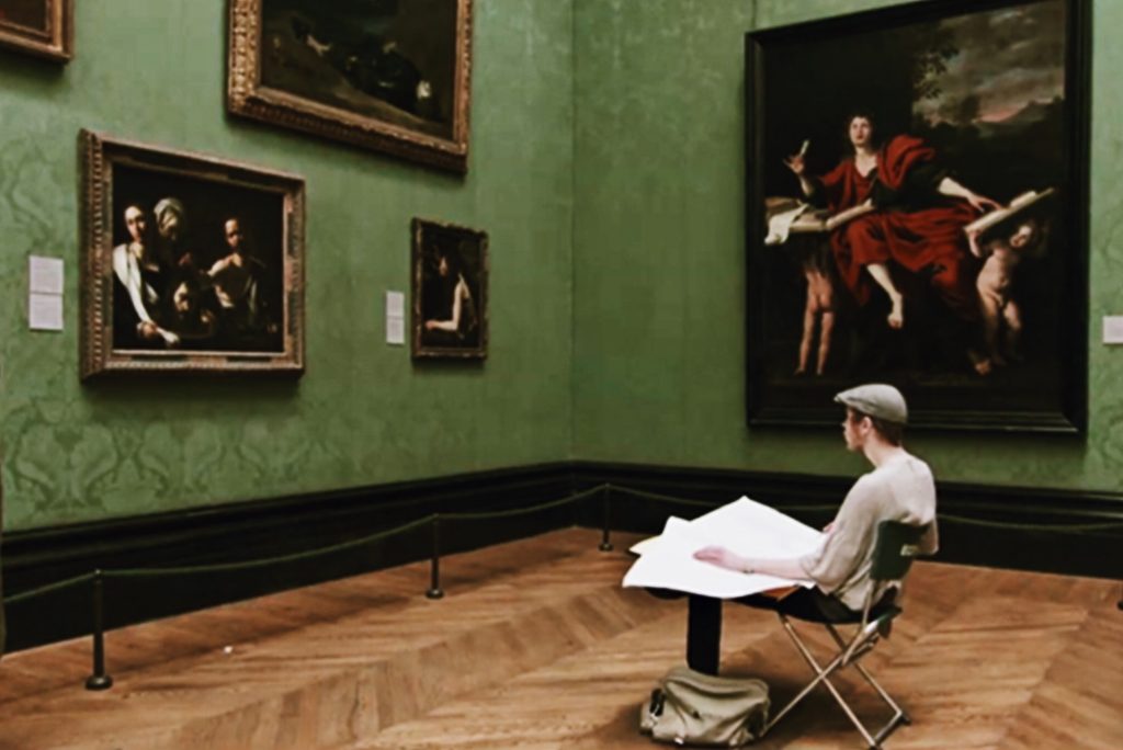 A person sketching with a large pad of paper on an easel while seated in a museum. They are surrounded by classical paintings in ornate frames hung on green walls. The artist is wearing a cap and white shirt, with a satchel on the floor.
