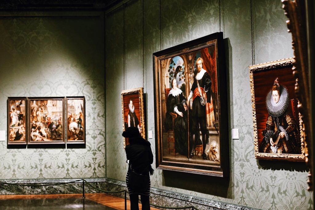 A person stands in a dimly lit art gallery, gazing at large framed paintings on the patterned walls. The ornate artworks depict historical figures, and the floor is polished wood. The atmosphere is quiet and contemplative.