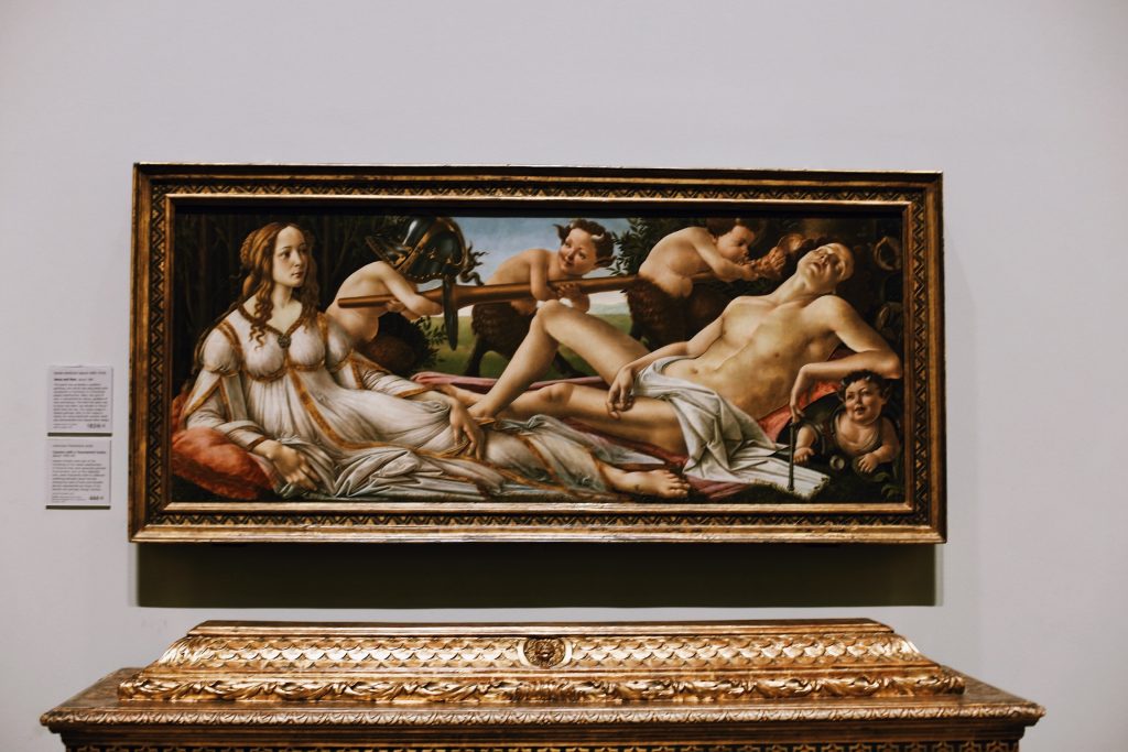 A framed classical painting featuring a reclining woman and several other figures in a detailed, natural setting. The artwork is displayed on a gallery wall with an ornate gold frame and a placard to the left.