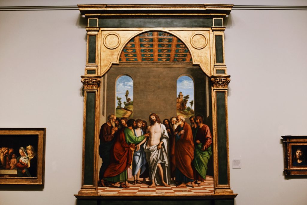 A classical painting of a biblical scene featuring Jesus surrounded by twelve disciples under an ornate archway. The background includes a landscape with trees and buildings, typical of Renaissance art. The painting is displayed in a museum.