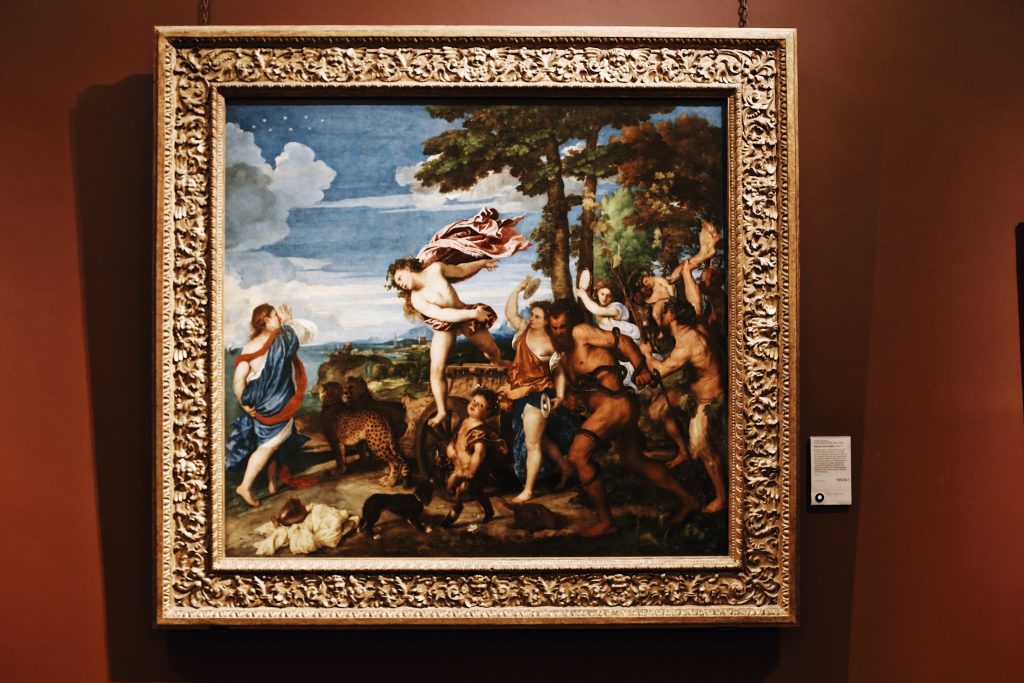 A framed classical painting depicting a mythological scene with a group of figures, including partially clothed men and women, some with musical instruments, dancing in a lush landscape with trees and a cloudy sky.