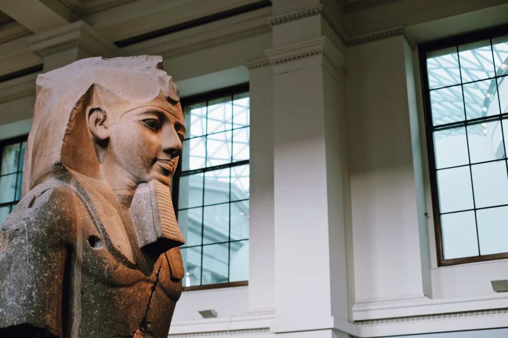 A large, ancient Egyptian stone statue of a pharaoh with a broken arm is displayed in a museum. The background features large windows with a grid pattern and a high ceiling. The lighting is soft and natural.