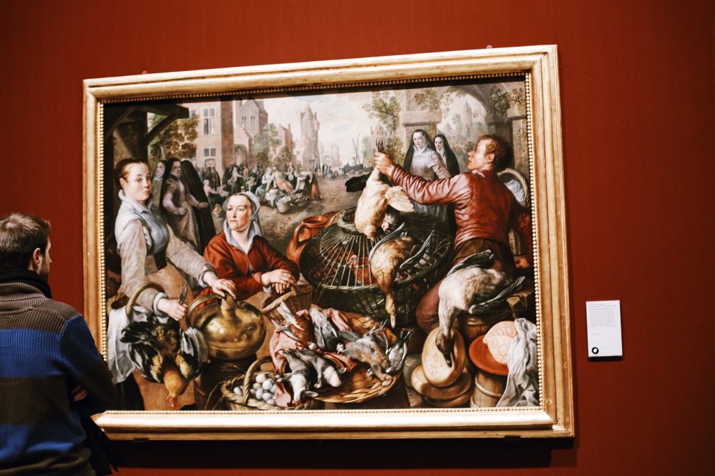 A person views a detailed painting in a museum. The artwork depicts a lively market scene with people handling poultry and various foods. The painting is framed in gold and displayed against a red wall.