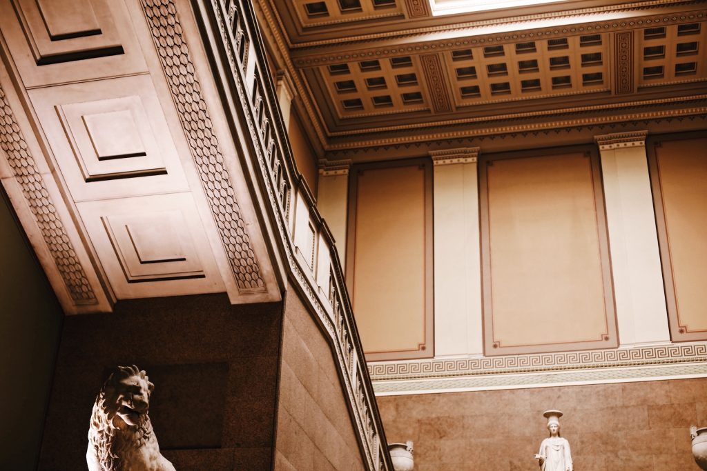 Interior of a grand building showcasing ornate architectural details. A stone lion sculpture is partially visible in the foreground, with a classical statue and intricately designed ceiling and walls in the background.
