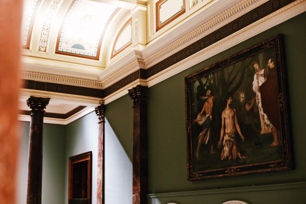 An ornate room features a high ceiling with intricate details and large dark columns. A classical painting depicting several figures is mounted on the green wall. The space is elegantly lit, creating a warm atmosphere.
