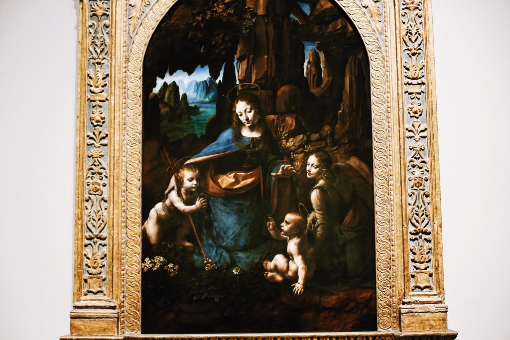 A Renaissance painting depicting a woman in blue with a halo, surrounded by three children in a lush landscape. The ornate gold frame features intricate carvings. The scene conveys a sense of tranquility and reverence.