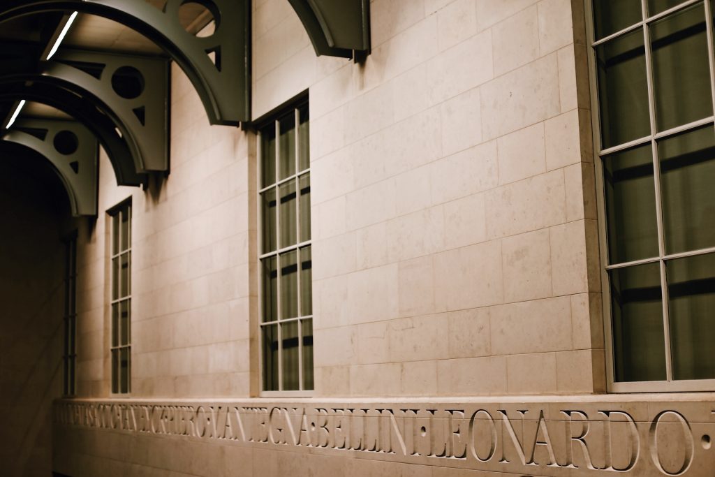 Interior hallway with large windows and arched ceiling beams. The stone wall features an engraved line of text in Italian, including the name Leonardo. The lighting is warm, highlighting the architectural details.