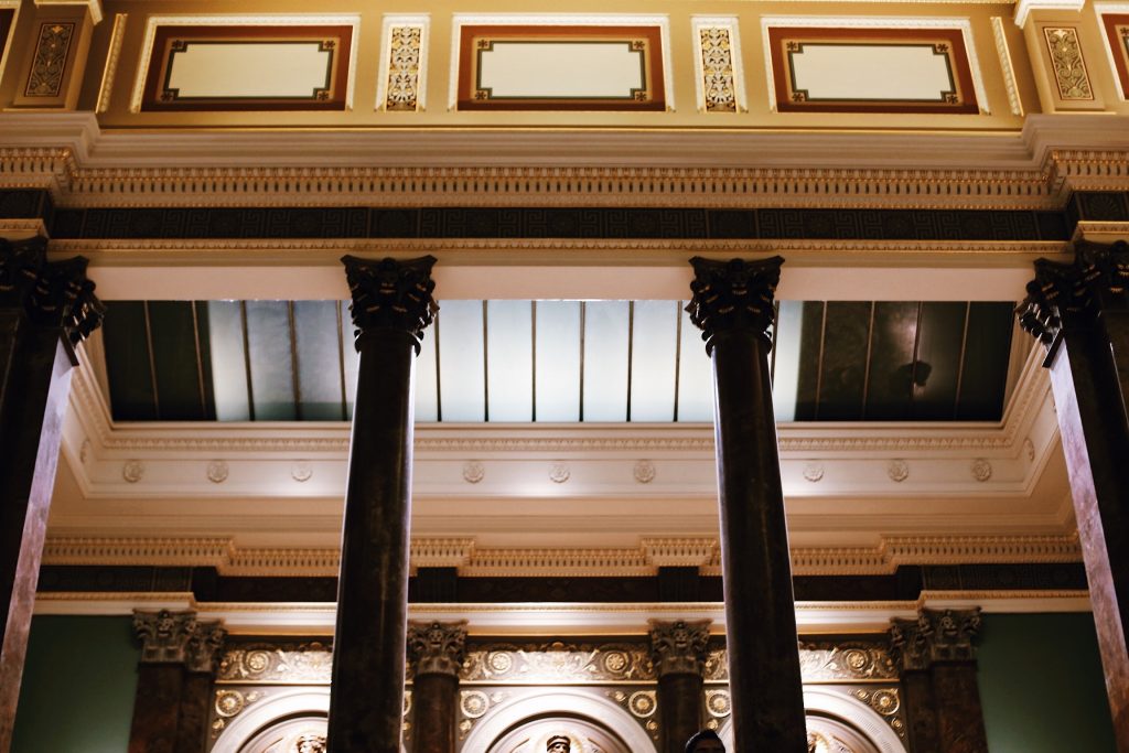 Interior view of a grand building with ornate architectural details, featuring tall, dark marble columns and an elaborately decorated ceiling with intricate moldings and panel designs. Warm lighting enhances the luxurious atmosphere.