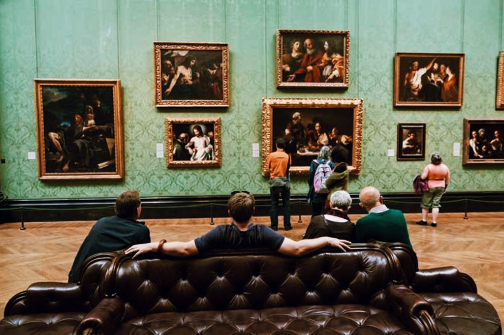 People sit on a brown leather sofa and stand in front of a wall displaying various framed paintings in an art gallery. The room has a green patterned wall and wooden floor, creating a classic and elegant atmosphere.
