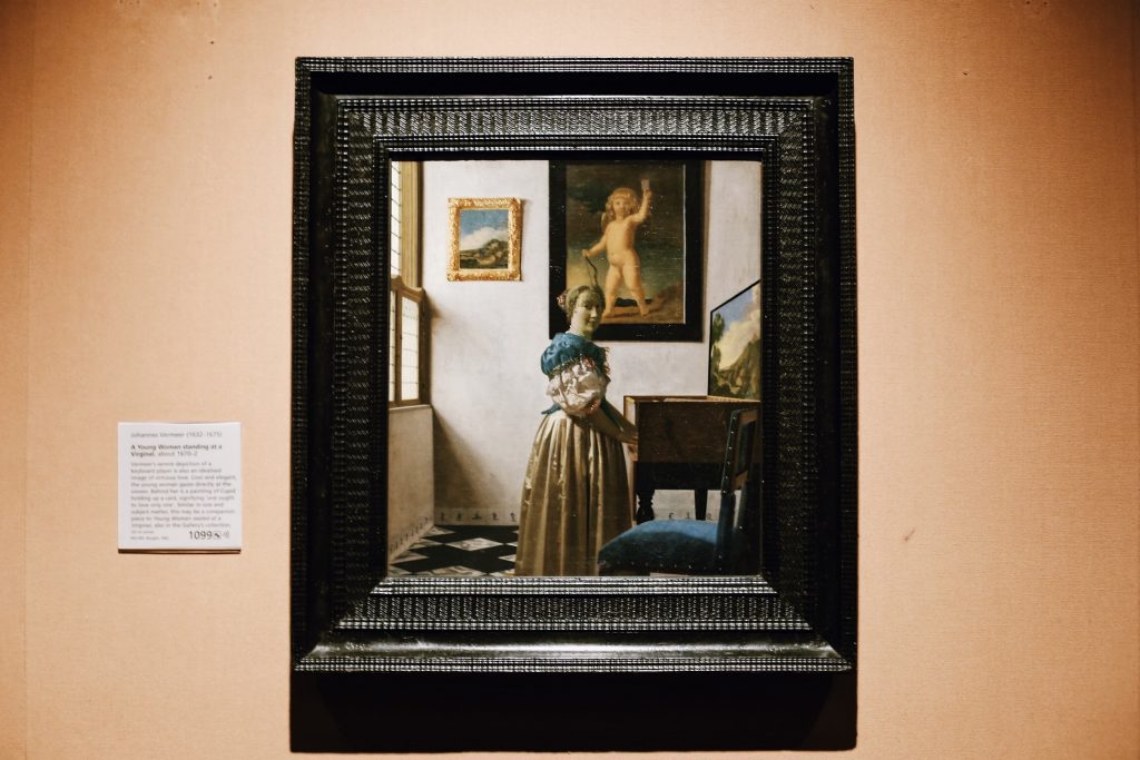 A framed painting depicting a woman standing in a sunlit room, reading a letter. The room features a window, a chair, and a painting on the wall. The frame hangs on a beige wall with an exhibit description beside it.