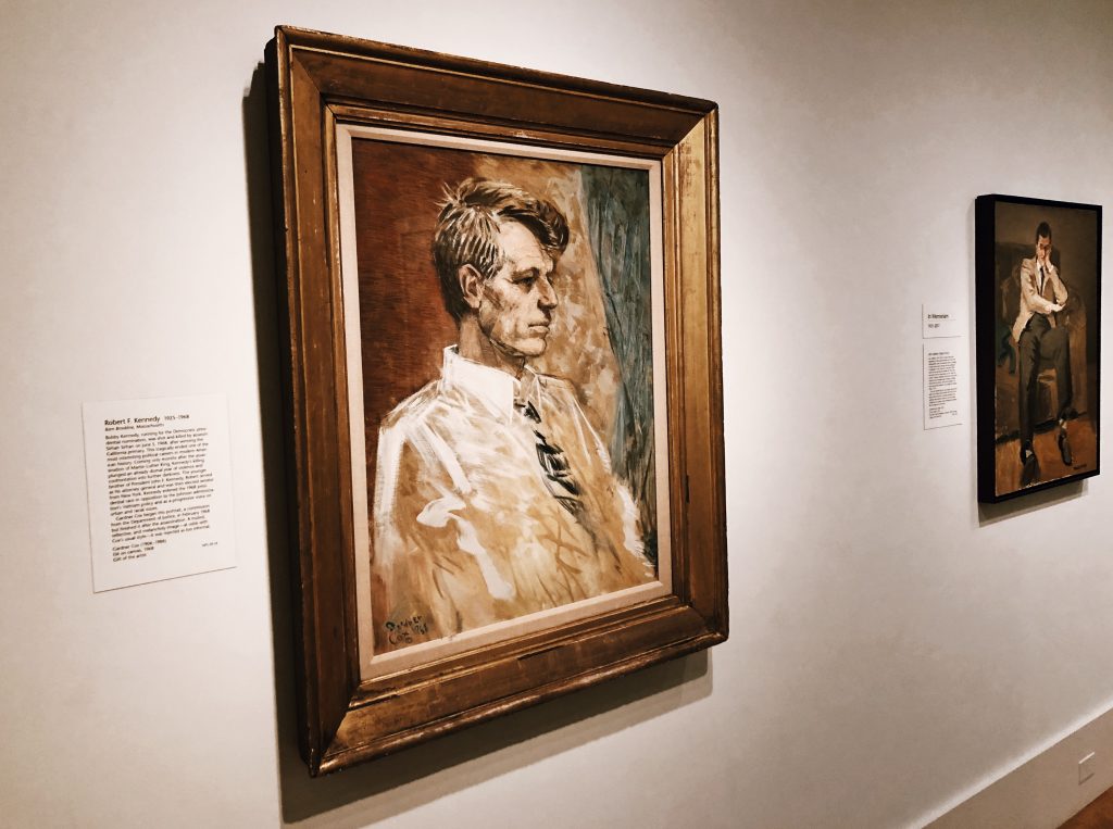 A museum wall displays a portrait of a man in a suit. The framed painting has a plaque with descriptive text next to it. Another, smaller painting hangs in the background, also with an accompanying plaque.