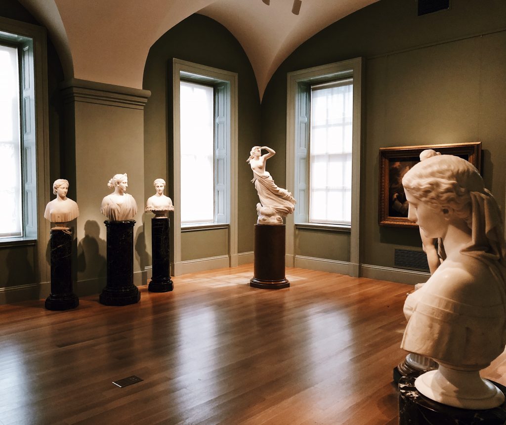 A museum room with several marble statues on pedestals, including busts against a wall and a full-body sculpture in the center. The walls are green, and natural light streams in through tall windows. A framed painting hangs on the right wall.