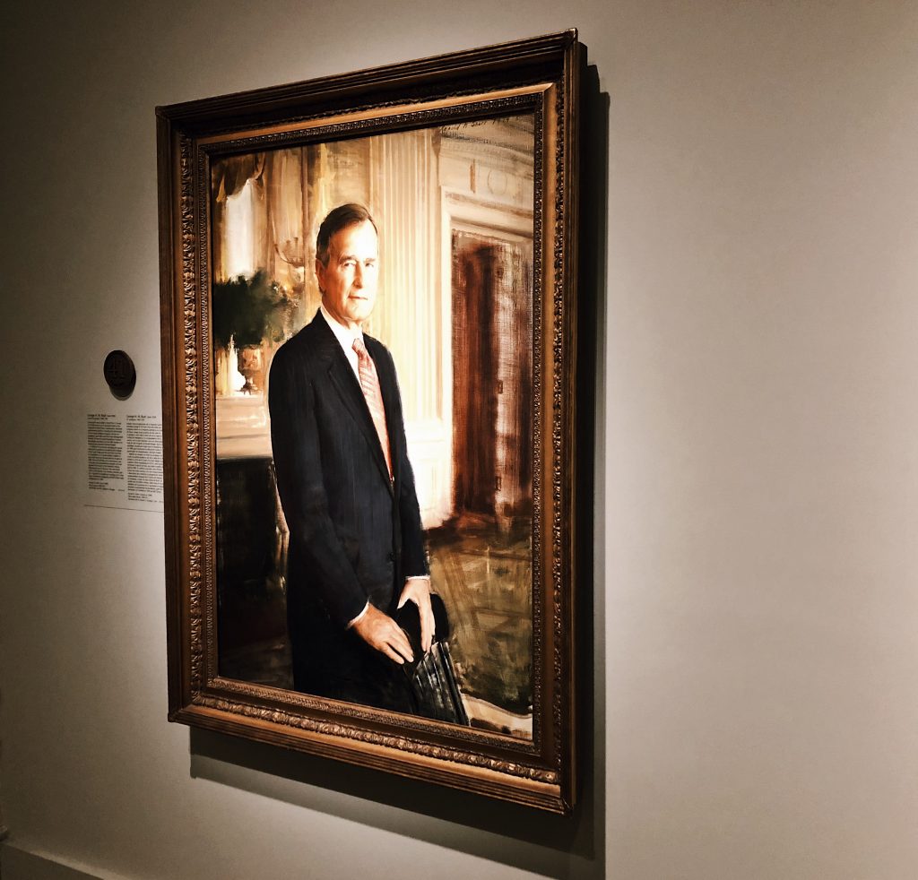 A framed portrait of a person in a suit, standing in a formal room with ornate decor. The lighting highlights the details of the painting and its gold frame. A plaque is mounted on the wall beside the portrait.