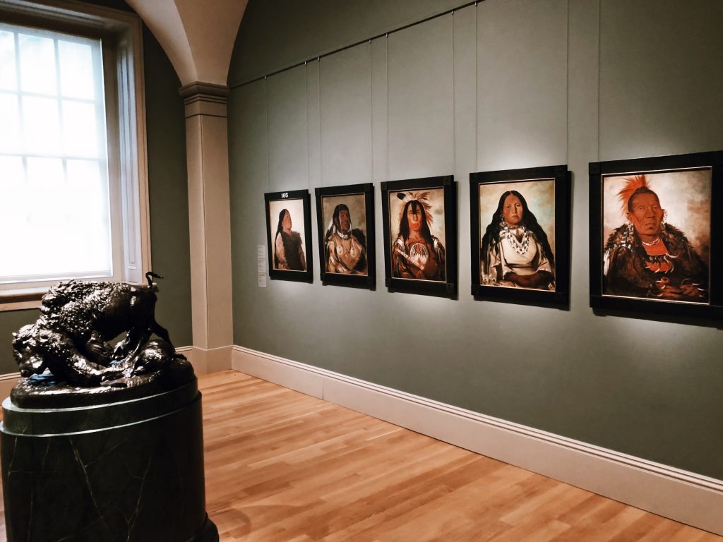Art gallery interior with five framed portraits of Indigenous individuals displayed on a pale green wall. A bronze sculpture of a peacock is positioned on a pedestal nearby. A large window allows natural light into the room.