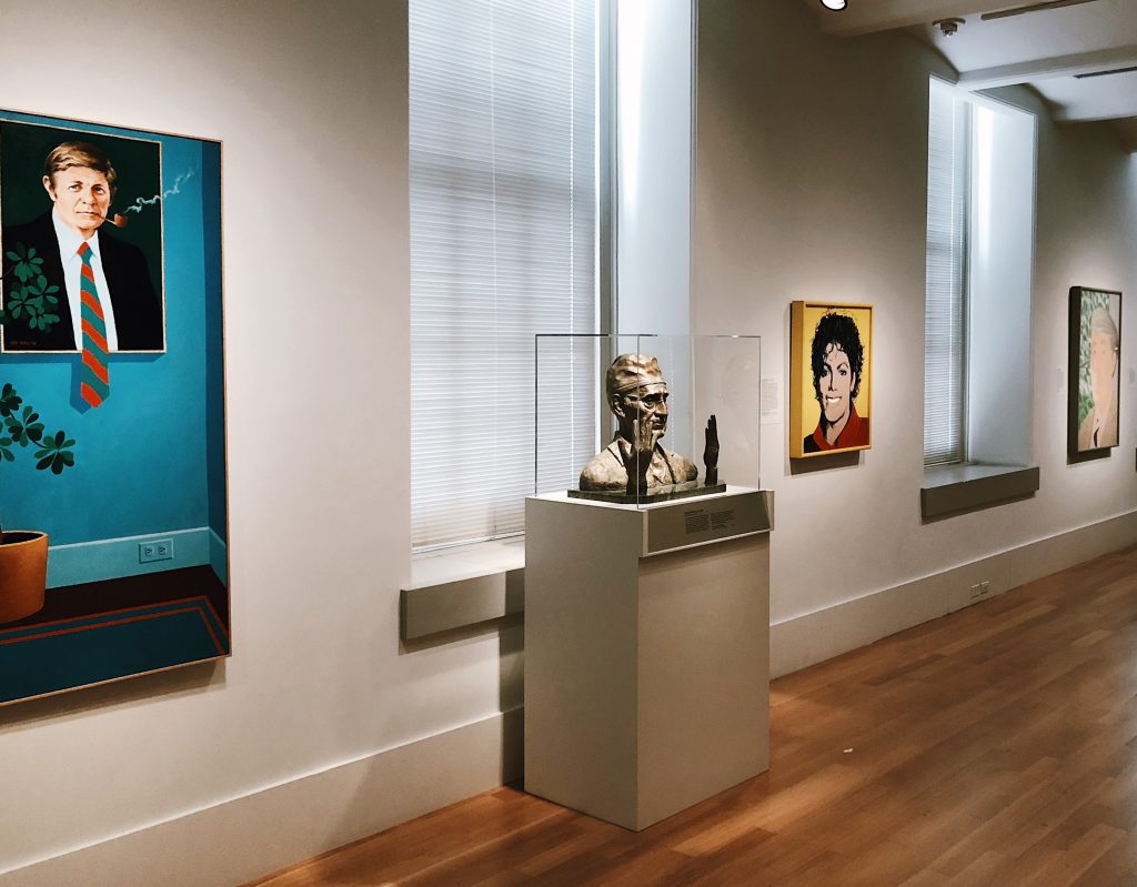 An art gallery with portrait paintings hanging on the walls and a sculpted bust displayed in a glass case. The artworks are in a well-lit room with wooden floors and large windows.
