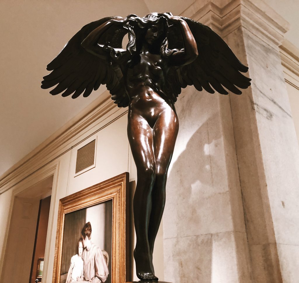 A bronze statue of an angelic figure with large wings stands with arms raised, positioned in an indoor setting. A framed portrait is visible on the wall in the background.